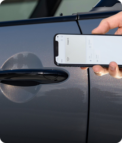 A hand holding up a smartphone open to the MINI App with a steering wheel in the background.
