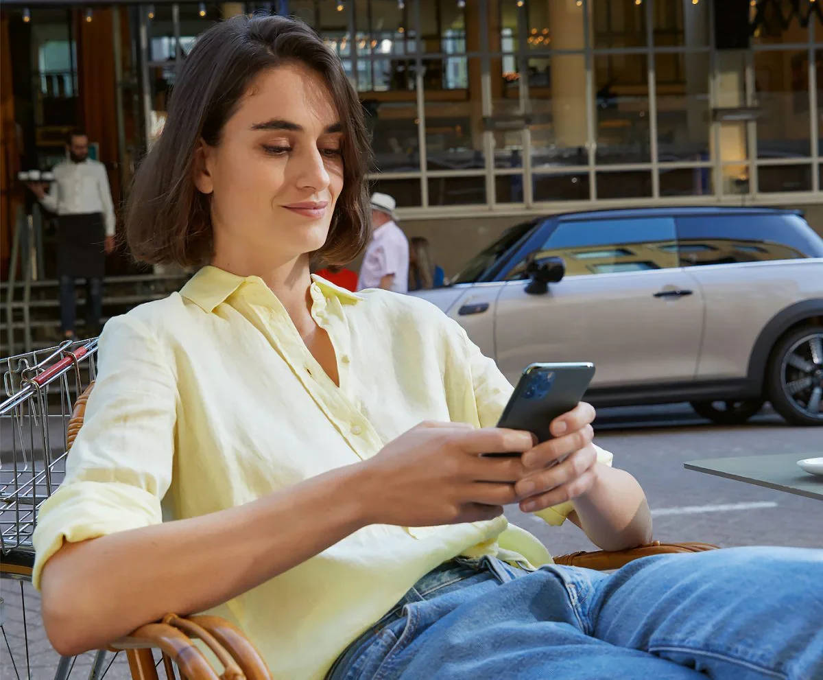 Female wearing yellow collared button-down shirt and jeans, sitting in a chair and holding a smartphone next to a street with a grey MINI vehicle parked in the background. | Flow MINI in Winston-Salem NC